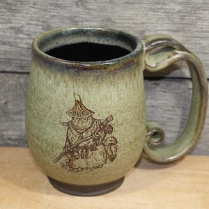 May include: A ceramic mug with a mottled sage green glaze and a dark brown rim. The mug features a brown etched illustration of a cloaked figure wearing a pointed hat and carrying a staff, standing next to a small mushroom.