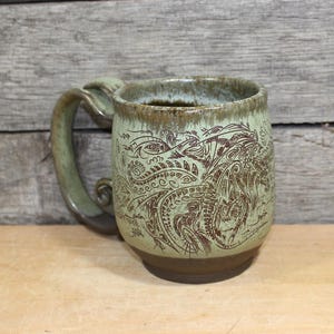 May include: A handmade ceramic mug with a unique design. The mug features a light green glaze with brown etched artwork. The handle is a matching light green color. The mug is sitting on a wooden surface.