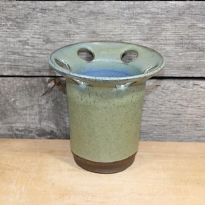 K&ouml;nnte beinhalten: Ein handgefertigter Zahnb&uuml;rstenhalter aus Keramik mit einer gesprenkelten gr&uuml;nen Glasur und einem braunen Boden. Die Oberseite hat zwei L&ouml;cher und einen blauen Innenraum. Der Halter ist zylindrisch und steht auf einer Holzoberfl&auml;che.