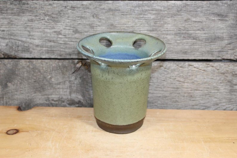 K&ouml;nnte beinhalten: Ein handgefertigter Zahnb&uuml;rstenhalter aus Keramik mit einer gesprenkelten gr&uuml;nen Glasur und einem braunen Boden. Die Oberseite hat zwei L&ouml;cher und einen blauen Innenraum. Der Halter ist zylindrisch und steht auf einer Holzoberfl&auml;che.