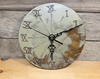 May include: A round wall clock with a mottled light green and brown surface. The clock face features black Roman numerals and intricate black metal hands. Thin black lines extend from the center to each numeral, decorated with small circular accents.
