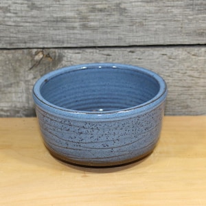 May include: A handmade, round, blue ceramic bowl with a textured surface and a slightly rough, unglazed base. The bowl has a simple, minimalist design, perfect for serving food or as a decorative piece. The bowl's diameter is approximately 6 inches.