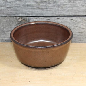 May include: A brown ceramic bowl with a dark brown rim. The bowl is round and has a smooth, glossy interior. The exterior has a textured appearance. The bowl is sitting on a wooden surface.