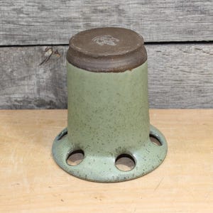 K&ouml;nnte beinhalten: Eine gesprenkelte, hellgr&uuml;ne Keramikvase mit braunem Rand und Boden. Die Vase hat eine zylindrische Form mit einem ausgestellten Boden mit mehreren kreisf&ouml;rmigen &Ouml;ffnungen. Die Vase steht auf einer Holzoberfl&auml;che.