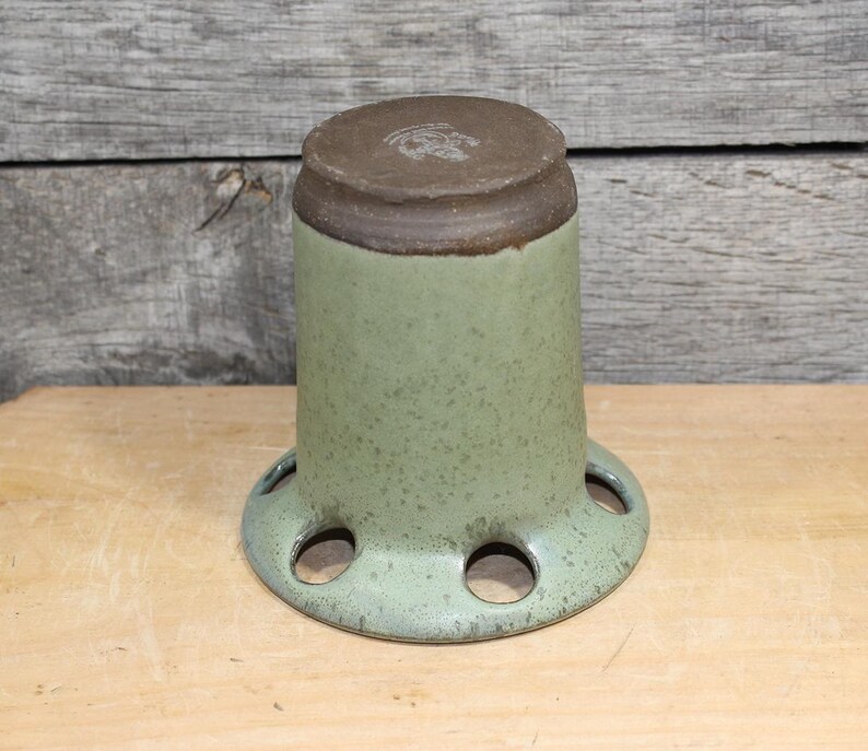 K&ouml;nnte beinhalten: Eine gesprenkelte, hellgr&uuml;ne Keramikvase mit braunem Rand und Boden. Die Vase hat eine zylindrische Form mit einem ausgestellten Boden mit mehreren kreisf&ouml;rmigen &Ouml;ffnungen. Die Vase steht auf einer Holzoberfl&auml;che.