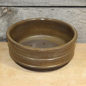 May include: A round, brown ceramic bowl with a textured surface and horizontal grooves. The bowl is set on a wooden surface, with a wooden background. The bowl is likely handmade and suitable for serving or display.