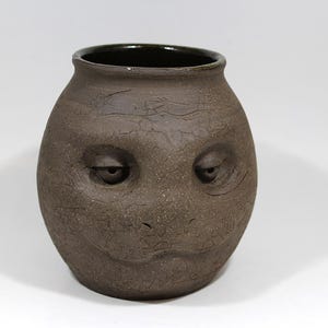 May include: A brown ceramic vase with a face sculpted on the front. The face has two eyes and a mouth.