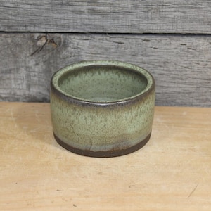 May include: A small, handmade ceramic bowl with a cylindrical shape. The bowl features a light green glaze with brown accents around the rim and base. The bowl is displayed on a wooden surface.