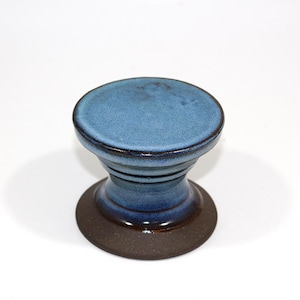 May include: A handmade ceramic bottle stopper with a blue glazed top and a dark brown base. The stopper has a unique hourglass shape and is suitable for wine bottles.