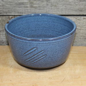May include: A handmade, blue ceramic bowl with a speckled finish. The bowl has a rounded shape and features three diagonal, brown etched lines on the side. The bowl is sitting on a wooden surface.