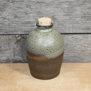 May include: A ceramic jug with a rounded body and a narrow neck, topped with a light brown cork stopper. The upper portion of the jug features a speckled light green and grey glaze, while the lower portion is a solid, dark matte brown finish.