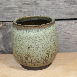 May include: A handmade ceramic tumbler with a speckled, light green glaze. The cup features a dark brown rim and base, with a delicate etched design of plants around the lower portion. The interior is a dark brown color.