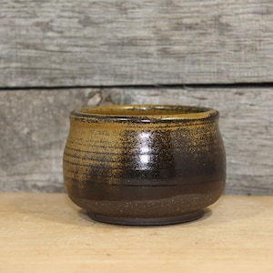 May include: A small, handcrafted ceramic bowl with a rounded shape. The bowl features a gradient of colors, transitioning from a light brown at the top to a dark brown at the base. Horizontal lines encircle the bowl, adding texture.