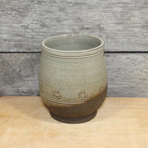May include: A cylindrical ceramic tumbler featuring a two-tone glaze with a light grey upper section and a dark brown textured base. The exterior is decorated with a repeating embossed circular cross pattern encircling the middle of the vessel.