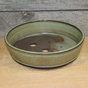 May include: A round, shallow ceramic bonsai pot with a speckled light green glaze on the exterior and rim. The interior base is dark brown and features two circular drainage holes. The pot has a smooth, slightly tapered silhouette and a flat bottom.