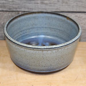 May include: A round ceramic bowl with a speckled light gray and blue glaze. The exterior features a textured, mottled finish, while the interior transitions into a deeper blue tone at the base. The rim is defined by a dark, thin, circular edge.
