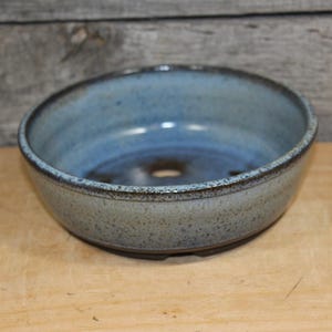 May include: A round ceramic bonsai pot with a speckled light blue and grey glaze. The interior features a deep blue hue with a visible drainage hole at the center of the base. The rim is dark and textured, contrasting with the lighter body.