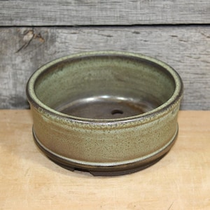 May include: A round, ceramic bonsai pot with a muted green glaze. The pot has a low profile with a wide opening and a drainage hole. The rim and base are a darker brown. The pot sits on a wooden surface.
