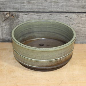 May include: A round ceramic bowl with a sage green glazed exterior and a dark brown interior. The bowl features a horizontal ridge detail around the middle of the outer wall and a small drainage hole centered at the bottom of the basin.