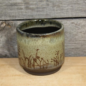 May include: A cylindrical ceramic cup featuring a mottled sage green and brown glaze with a dark interior. The exterior surface is decorated with etched brown line drawings of various wild grasses, ferns, and small plants encircling the base.