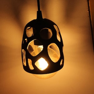 May include: A black pendant light fixture with an irregular, organic cutout pattern across its surface. The shade features various circular and oval openings that allow a warm, glowing light to shine through from the internal light bulb.