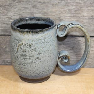May include: A handmade ceramic mug with a unique handle design. The mug features a light blue and gray speckled glaze with a dark rim and a decorative illustration. The handle is a light blue color with a curled design.