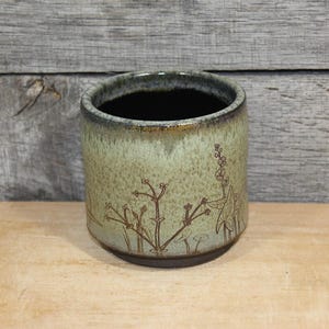 May include: A cylindrical ceramic cup featuring a speckled sage green glaze with a dark brown rim and base. The exterior is decorated with etched brown line drawings of wildflowers and stems, while the interior is finished in a deep black glaze.