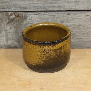 May include: A small, round ceramic container with a textured, speckled surface. The exterior transitions from a dark brown base to a lighter, golden-brown upper portion. The rim is a dark brown color.