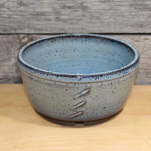 May include: A handmade ceramic bowl with a speckled, light blue exterior and a darker blue rim. The bowl features three decorative, carved lines on the side. The interior is a solid light blue. The bowl sits on a dark brown base.