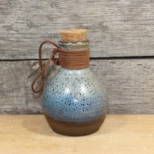 May include: A small, round ceramic bottle with a cork stopper and a brown leather strap. The bottle has a speckled blue and brown glaze. The leather strap is wrapped around the neck of the bottle.