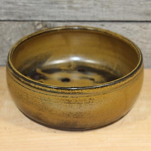 May include: A round, ceramic bowl with a wide rim. The bowl has a dark brown interior and a golden-brown exterior with dark accents. The bowl is likely handmade and suitable for serving food or as a decorative piece.