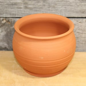 May include: A round, unglazed terracotta pot with a wide opening. The pot has a slightly bulbous shape with horizontal ridges around the body. The color is a warm, earthy orange-brown. Ideal for planting flowers or herbs.