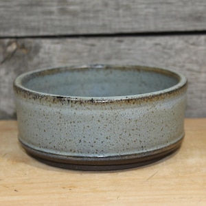 May include: A round ceramic bowl with a speckled light blue and gray glaze. The bowl features a dark brown rim and a matching dark brown base, with a smooth, cylindrical wall and a wide, open top design for holding various items.