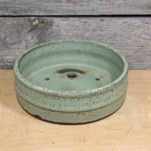 May include: A round, handmade ceramic planter in a muted green hue. The planter has a speckled texture and a dark brown rim. It features a drainage hole and two smaller holes. The planter sits on a wooden surface.