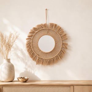 Peut inclure: Un miroir mural circulaire doté d'un cadre en corde macramé beige. Le miroir présente une vitre centrale entourée de motifs de cordes nouées complexes et d'une frange de fils pendants. Une boucle de corde unique dépasse du sommet.
