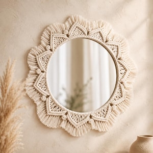 Peut inclure: Un miroir mural circulaire encadré par une bordure en macramé blanc cassé complexe. La bordure décorative présente un motif géométrique répétitif de cordons noués et de franges, créant un bord festonné en forme de pétale autour du miroir central.