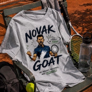 May include: A sport grey t-shirt featuring a graphic illustration of a tennis player with the text NOVAK GOAT, YOU CANNOT BE SERIOUS, and HE BREAKS RECORDS AND OPPONENTS. The design includes a tennis racket and a crown icon in dark blue and green.