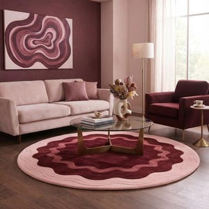 Puede incluir: Una alfombra circular grande con bandas onduladas concéntricas en tonos rosa claro, rosa viejo y burdeos intenso. La alfombra tiene una superficie texturizada y copetuda, colocada sobre un suelo de madera oscura, a juego con el arte abstracto mural.