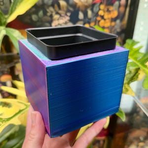 May include: A square, 3D-printed planter with a gradient finish transitioning from blue to purple. The planter features a smooth, layered texture and includes a removable black plastic square insert tray resting on the top opening of the container.