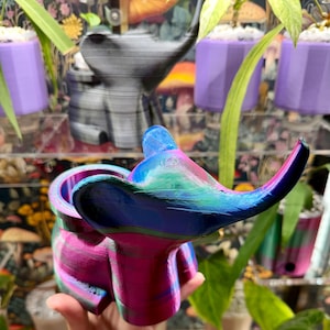 May include: A 3D printed elephant-shaped planter featuring a vibrant, multi-colored gradient finish that transitions between shades of magenta, blue, green, and purple. The planter has a smooth, curved form with a hollow opening at the top.