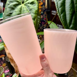 May include: Two pale pink glass tumblers with a smooth, matte finish. The taller tumbler is cylindrical with a flat rim, while the shorter tumbler features a subtle, spiraling ribbed texture along its exterior surface and a rounded base.