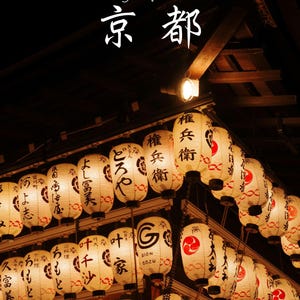 以下が含まれることがあります： 暗い木造建築から吊るされた、明かりが灯る白い提灯のコレクション。各提灯には黒い日本語の書道と赤い円形の模様が描かれています。上部には「Kyoto」という文字と「京都」という漢字が表示されています。
