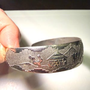 May include: A silver-toned metal cuff bracelet featuring an embossed landscape scene. The design includes a small house, a cactus, and a mountain silhouette against a textured background. The bracelet has an open back design and a polished finish.
