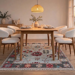 Op de afbeelding: Een rechthoekige houten eettafel met vier stoelen met witte, gestructureerde bekleding en houten poten. De tafel is gedekt met borden en een vaas, geplaatst op een lichtblauw vloerkleed met een donkergrijze rand en bloemmotieven.