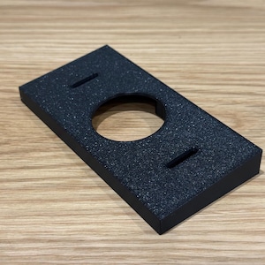 May include: A rectangular black mounting plate with a textured finish, featuring a large circular cutout in the centre and two smaller horizontal rectangular slots positioned above and below the central opening for secure installation purposes.