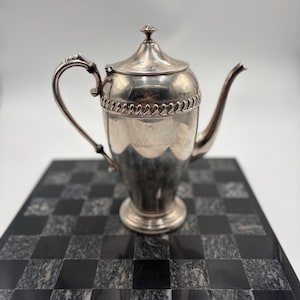 May include: A silver-toned metal teapot featuring a tapered body, a curved handle, a slender spout, and a removable lid with a small finial. The upper section of the teapot is decorated with a textured rope-like band encircling the circumference.