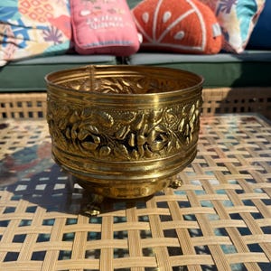Puede incluir: Una maceta redonda de metal en tono dorado con un intrincado patrón floral y de hojas en relieve alrededor de su exterior. El recipiente se apoya sobre tres pequeños pies decorativos y tiene una parte superior abierta y brillante.