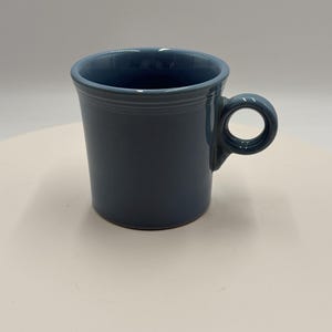 May include: A solid slate blue ceramic mug with a smooth, glossy finish. The mug features a cylindrical body with a slightly flared rim and a circular handle attached to the side. The design includes subtle horizontal ridges near the top rim.