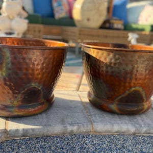 Peut inclure: Deux bols ronds assortis présentant une texture martelée et une finition multicolore avec des tons profonds de cuivre, de bronze et des reflets irisés verts et bleus. Chaque bol a une large ouverture et une base lisse et arrondie.