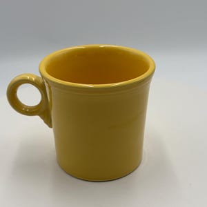 Könnte beinhalten: Ein leuchtend gelber Keramikbecher mit glatter, glänzender Oberfläche. Der Becher hat einen zylindrischen Körper mit einem leicht ausgestellten Rand und einen kreisförmigen Henkel an der Seite. Das Design ist schlicht und funktional.